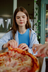 A young woman dressed casually sits at a table, pulling a slice of pizza from a large box. The atmosphere is relaxed, with greenery in the background and natural light streaming through the windows.