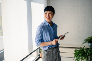 Asian woman in blue shirt holding a tablet and smiling indoors near large windows and plants, looking confident and approachable like a professional