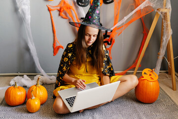 A person in a witch hat sits on the floor, using a laptop amid Halloween decorations and pumpkins, capturing the festive spirit