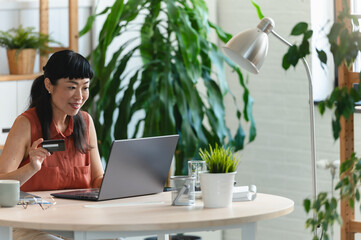 Smiling middle-aged Asian woman holding a credit card while shopping online on a laptop at home...
