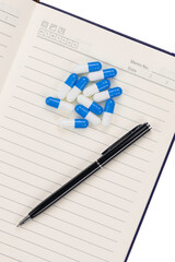 Notebook with blue and white capsule pills and black pen for medical schedule and planning.