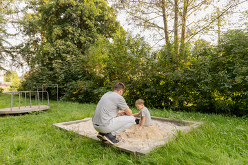 Fototapeta premium A father enjoys quality time with his young child at a playground. They are sitting in a sandbox, playing and laughing together, surrounded by lush nature on a sunny afternoon