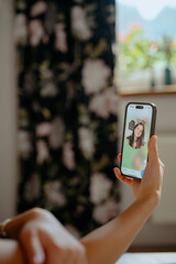 A person holds a smartphone displaying a smiling individual on a video call. The setting is cozy with floral curtains and soft light, creating a warm atmosphere during a casual interaction.