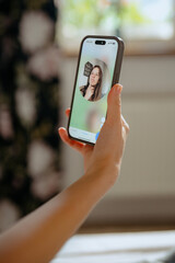 A woman is participating in a video call, holding her smartphone in one hand. She appears focused, with a cozy indoor setting visible in the background and natural light coming through the window.