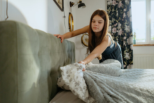 A young girl is making her bed in a bright bedroom filled with natural light. She carefully adjusts the sheets and blankets, showcasing her attention to detail and organization. - Powered by Adobe