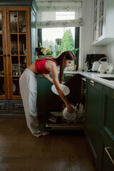 A woman in casual clothing is putting clean plates into a dishwasher in a contemporary kitchen. The tidy space features green cabinetry and bright natural light from the window.