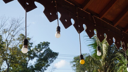 row of hanging light bulbs suspended from wooden roof with decorative trim, four white and one yellow bulb, set against blue sky, trees, and power lines in outdoor scene