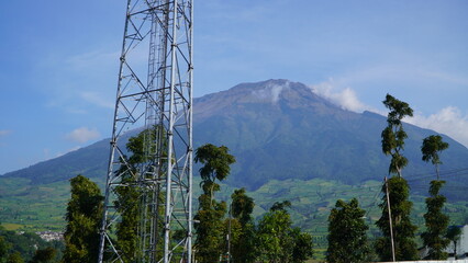 mountain landscape with cloud-covered peak&mdash;lush green base and rocky summit, tall metal tower and trees in foreground under clear sky with scattered clouds