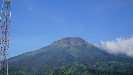 A towering, green-sloped volcano dominates the skyline beneath a clear blue sky, framed on the left by a tall transmission tower in a sunny outdoor landscape.