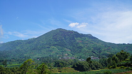 lush green mountain under clear blue sky&mdash;dense vegetation and small village at base, soft clouds and layered terrain create tranquil natural landscape