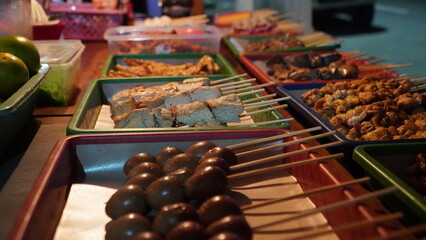 street food stall with skewered items including meat, tofu, and eggs neatly arranged in trays on table, outdoor market setting with background containers and vehicle