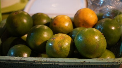 basket filled with green and partially yellow oranges, uneven coloration suggests mixed ripeness, placed on surface with plastic bag and white object in casual market or home setting