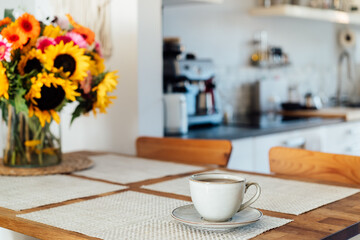 Focus on cup of coffee or tea and vase with huge sunflowers bouquet on wooden table in cozy modern interior of kitchen. Cozy Design home decor. summer mood, good morning routine. Selective focus.