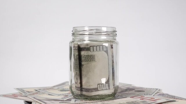 Rotating transparent glass jar filled with dollar bills, surrounded by scattered banknotes, symbolizing savings, finance, and wealth accumulation.