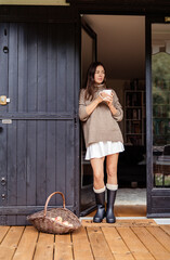 Young woman in rubber boots and cozy autumn outfit standing at rustic wooden door on terrace with cup of coffee. Relaxed outdoor countryside  lifestyle, slow living and downshifting  concept