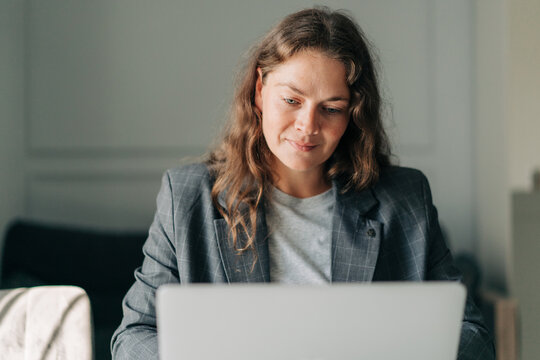 A middle-aged woman sitting at home in a business suit works on a laptop. - Powered by Adobe