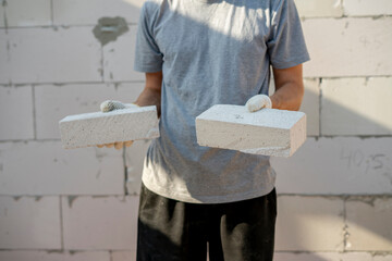 A construction worker in a gray shirt holds two bricks at a job site, focused on proper placement and structural integrity during construction activities.