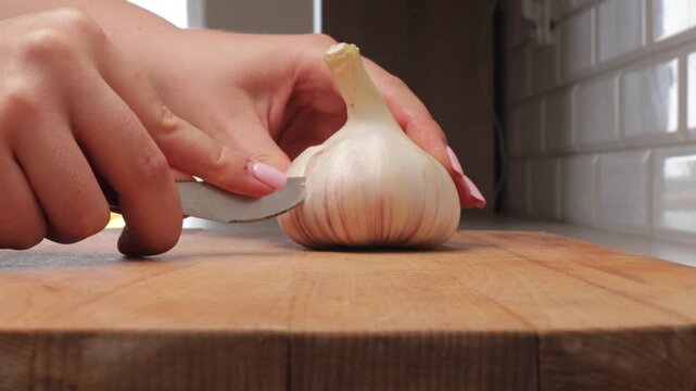 Peeling garlic on wooden cutting board. Close-up of hands using knife to clean fresh garlic for cooking preparation.