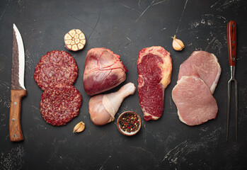 Selection of assorted raw meat cuts for zero carb carnivore diet: uncooked beef steak, pork, ground meat patty, heart, and chicken legs on black stone background top view with knife and carving fork