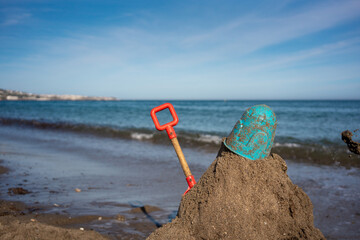 Red shovel and blue bucket placed on top of a sandcastle mound at the beach with calm ocean waves...