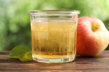 Delicious cider in glass, apple and green leaves on wooden table outdoors, closeup