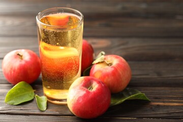 Delicious cider in glass, apples and green leaves on wooden table, closeup. Space for text