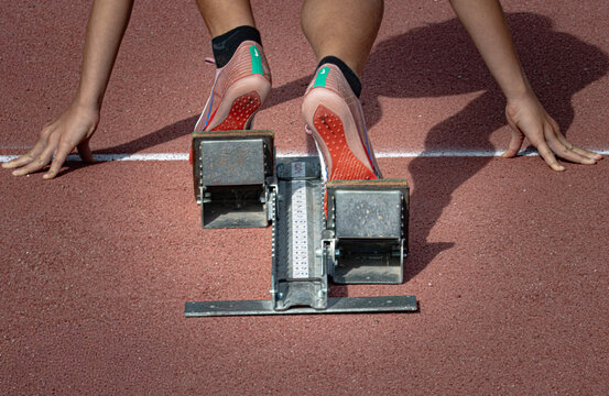 A runner crouched at the starting line of a track. His hands are planted on the ground, behind a white line. He stands in a starting block, his running shoes pressing against the inclined footrests.