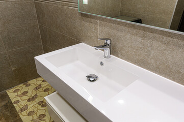 A modern bathroom sink with chrome faucet and drain. The countertop is white, and the walls are tiled in beige. A patterned rug is visible