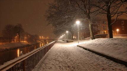 Snow falls gently over a quiet riverbank path, lit by warm street lamps. Trees line the walkway, creating a serene atmosphere on this cold winter evening.