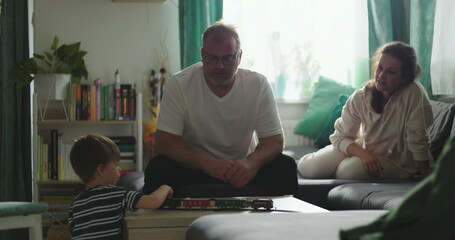 Father sitting with boy playing with toy train on coffee table, mother smiling and observing from...