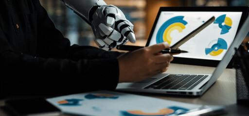 A close-up view of a human hand collaborating with a robotic hand over a laptop, analyzing data and charts in a modern office setting, showcasing advanced technology.Noogenesis