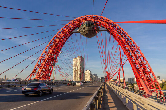 Zhivopisniy Bridge's striking red cable-stayed arch with glass observation capsule resembling a flying saucer over Moscow River. Modern architectural marvel under blue sky. - Powered by Adobe