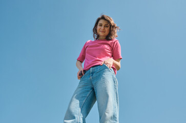 Beautiful carefree young woman with curly hair in pink t-shirt smiling and looking down at camera on blue sky background low angle shot.