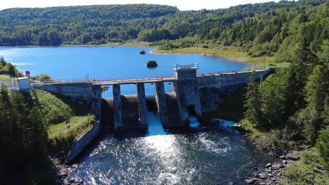 Aerial view moving away from an old concrete hydroelectric dam controlling the level of a forest lake. Mitis Dam, Mitis Lake, Quebec, Canada, 2025.