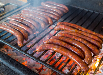 A numerous sausages sizzling on a grill over hot coals, with smoke rising around them. The sausages are brown and appear well-cooked, some with visible grill marks.