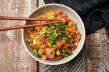 Delicious fried rice with vegetables in bowl and chopsticks on wooden table, top view