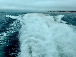 Dynamic boat wake carving through teal ocean under an overcast sky, strong sense of motion and coastal travel along the Cape Cod shoreline