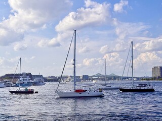 Sailboats and yachts anchored on a summer afternoon with fluffy cumulus clouds and a distant truss bridge spanning the harbor in Boston, MA