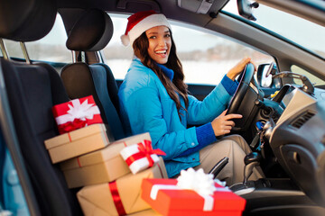 Happy woman in Santa hat driving car with pile of Christmas presents nearby, delivering gifts and enjoying winter holiday preparation