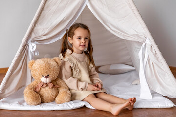 Happy 4-5 years old little girl in brown dress sitting in a wigwam tent with soft toy teddy bear. Children's room, childhood, games, imagination. High quality photo. Copy space © Irina Mikhailichenko