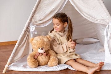 Happy 4-5 years old little girl in brown dress sitting in a wigwam tent with soft toy teddy bear and clapping her hands. Children's room, childhood, games, imagination. High quality photo. Copy space © Irina Mikhailichenko