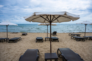Rows of empty lounge chairs and white umbrellas line a quiet, sandy beach under a cloudy sky, facing the calm sea.