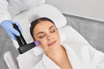 Brown haired woman receiving radiofrequency lifting procedure around eyes for her face skin rejuvenation at aesthetic cosmetology center