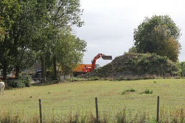 a crane is digging rubble and ground in a tipping trailer at a farm and a green meadow with trees in front in the countryside