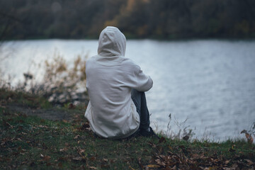 Lonely young man sits on the river bank in cloudy autumn weather, back view