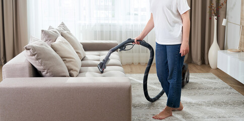 young woman in white shirt and jeans cleaning carpet under sofa with vacuum cleaner in living room,...