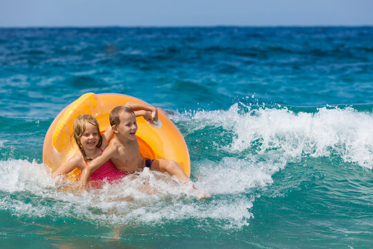 Happy kids have fun in sea surf on beach. Joyful couple of children on inflatable ring ride on breaking wave. Travel lifestyle, swimming activities in family summer camp. Vacations on tropical island