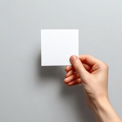 Obraz premium Elegant Hand Presenting a Blank White Square Card Mockup on a Minimalist Gray Background.