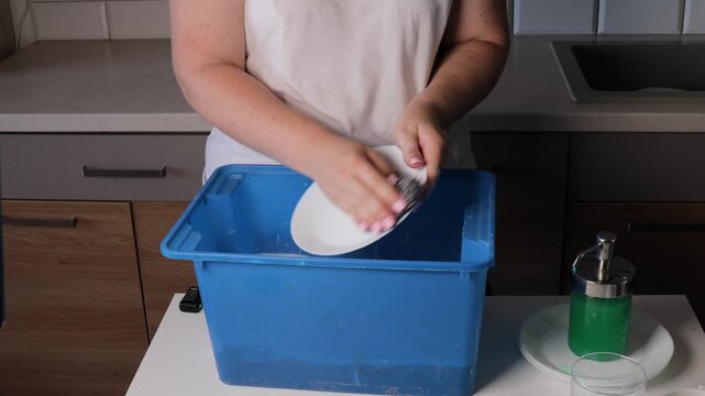 washing dishes in blue plastic basin without running water in home kitchen. woman using sponge to clean plate during water outage. concept of water saving, emergency routine, sustainable living