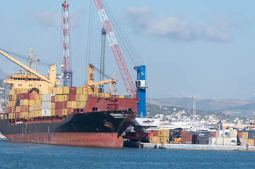 Naklejka premium Unloading large ocean container vessel, sea transport of containers, container shipping, ship charter, international transportation, port of Trapani, Sicily, Italy